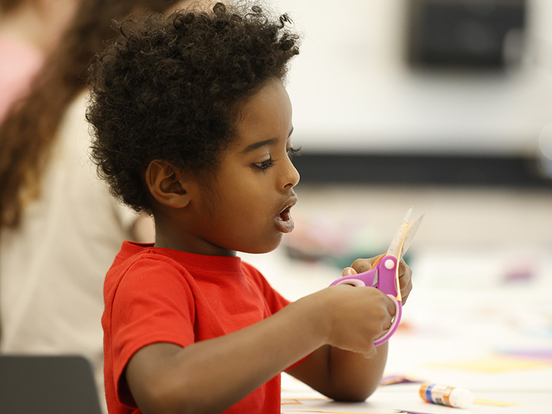 Photo of a young boy holding a pair of pink scissors while doing an art activity.