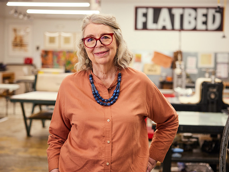 Photo of Katherine Brimberry in a printmaking studio with a sign that reads Flatbed on the wall behind her.