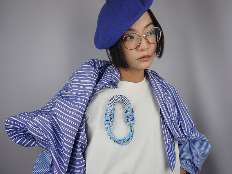 Photo of artist Dongyi Wu wearing a blue beret, blue pinstriped shirt, and a white T-shirt adorned with a handmade blue necklace.