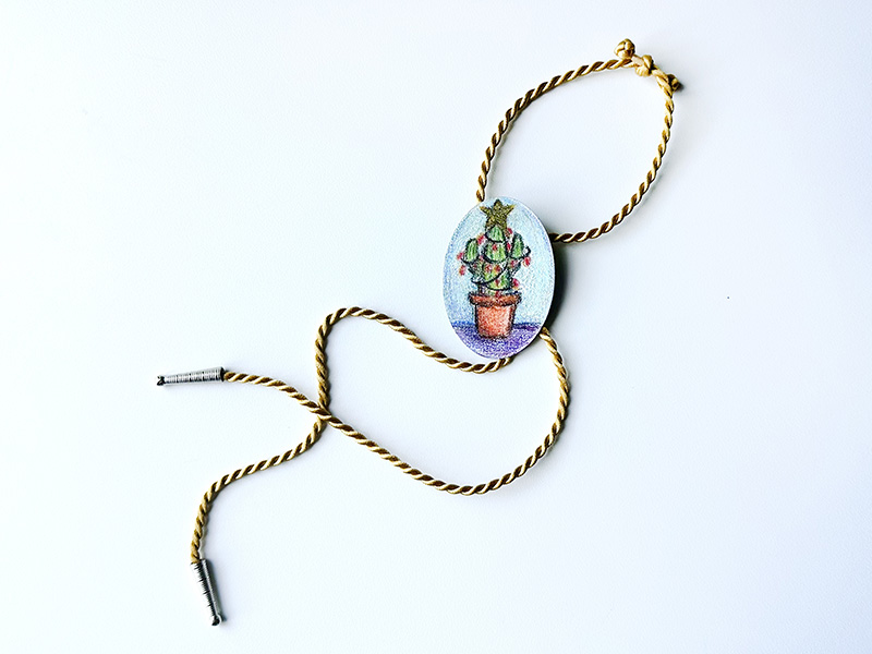 Photo of a golden rope bolo tie with an emblem featuring a drawing of a Christmas tree.