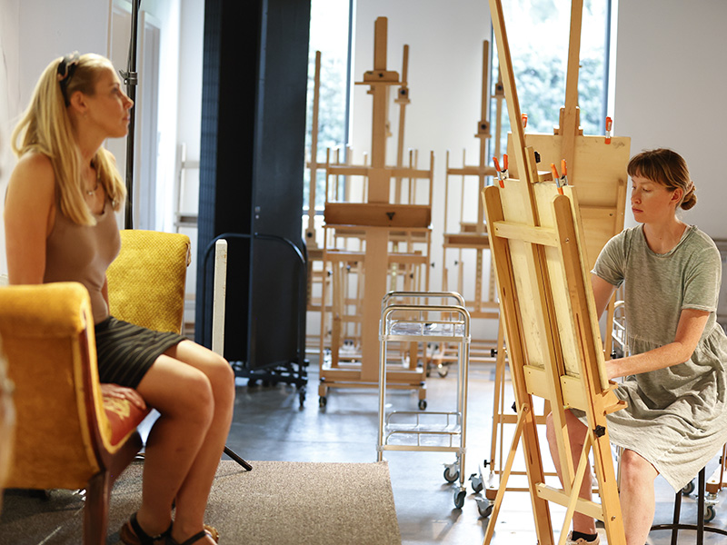 Photo of a woman sitting at an art easel drawing a portrait of another woman sitting opposite of her on a sofa.