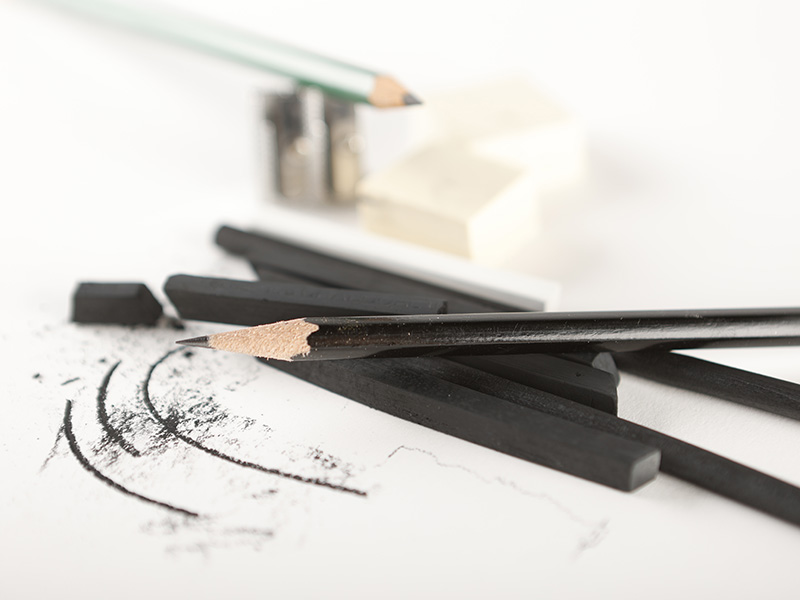 Photo of pencils, charcoals, a pencil sharpener, and two erasers on a white tabletop.