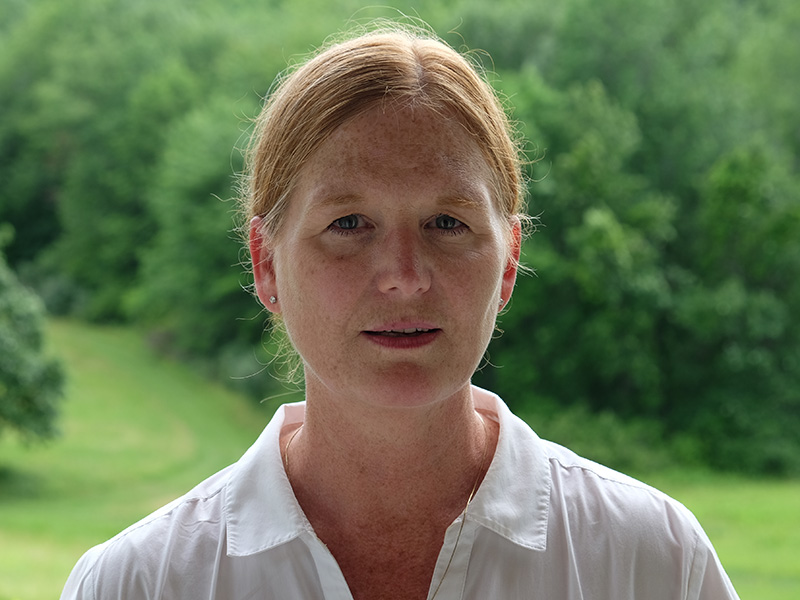 Photo of Anne Lindberg wearing a white shirt with her red hair pulled back standing in front a green meadow with trees.