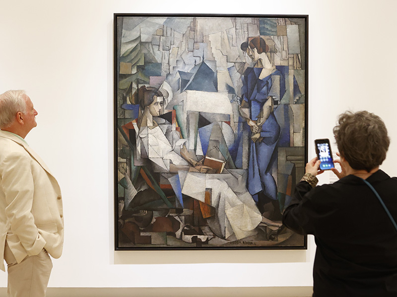 Photo of a man standing next to a large Cubist painting of two women in an art gallery while a woman next to him takes a photo of the painting on her cellphone.