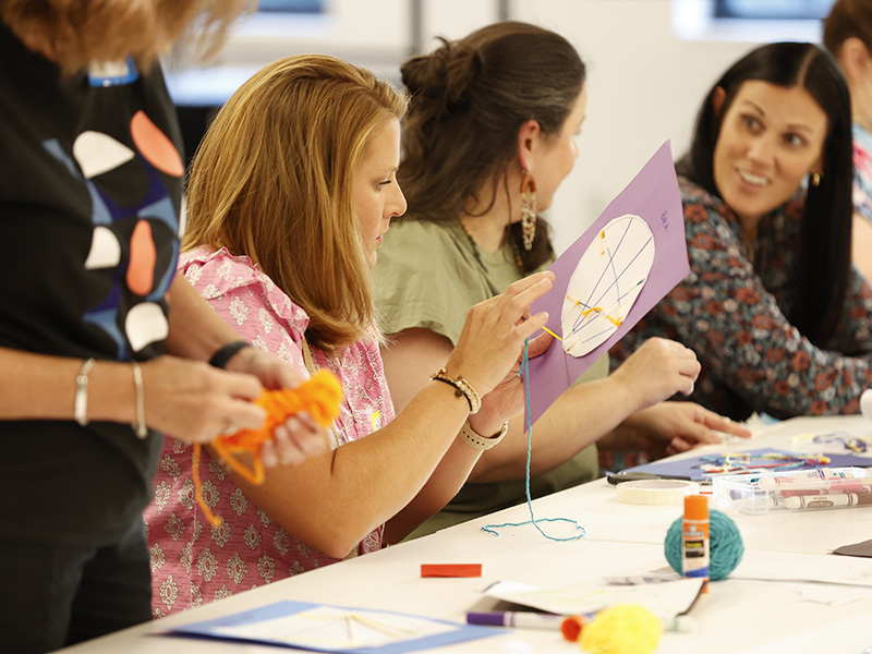 Photo of several white women sitting at a white table doing an art activity with paper, yarn, and glue sticks.