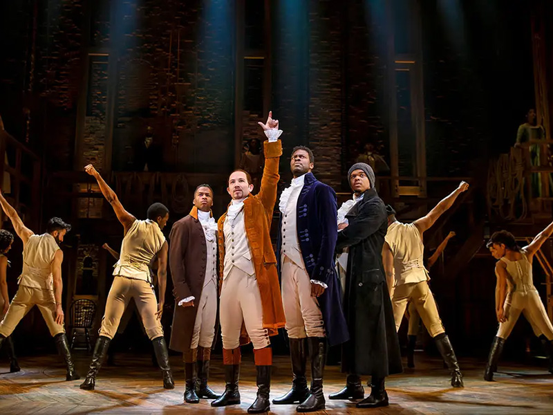 Photo of the Broadway cast of Hamilton performing on stage. Fergie Philippe stands on the far right of a group of four men in the foreground with four dancers posed in the background.