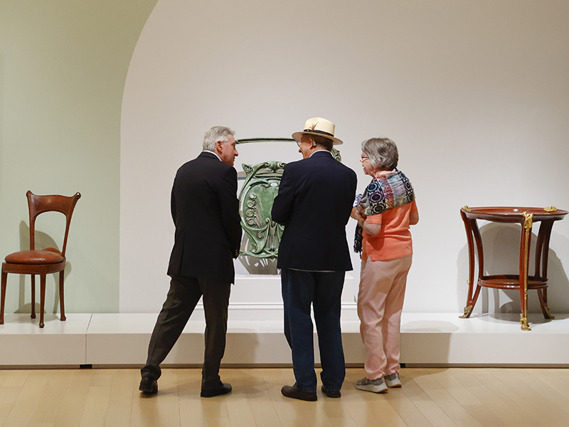 Photo of two men and one woman looking at Art Nouveau furniture in an art gallery.