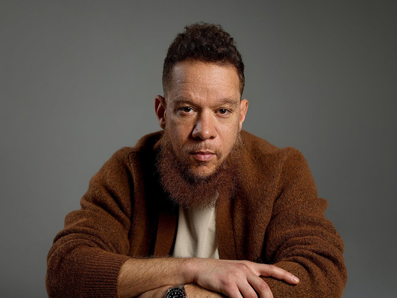 Headshot of Yashua Klos wearing a white shirt and brown sweater with his arms crossed in front of a gray background.