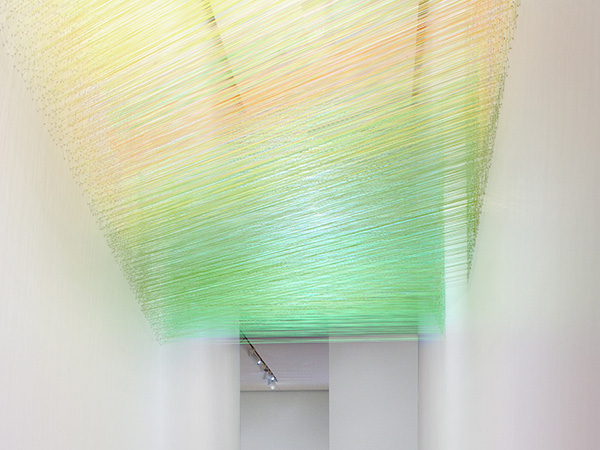 Photo of a multi-colored thread art installation by Anne Lindberg handing between two walls in a narrow corridor of an art gallery.