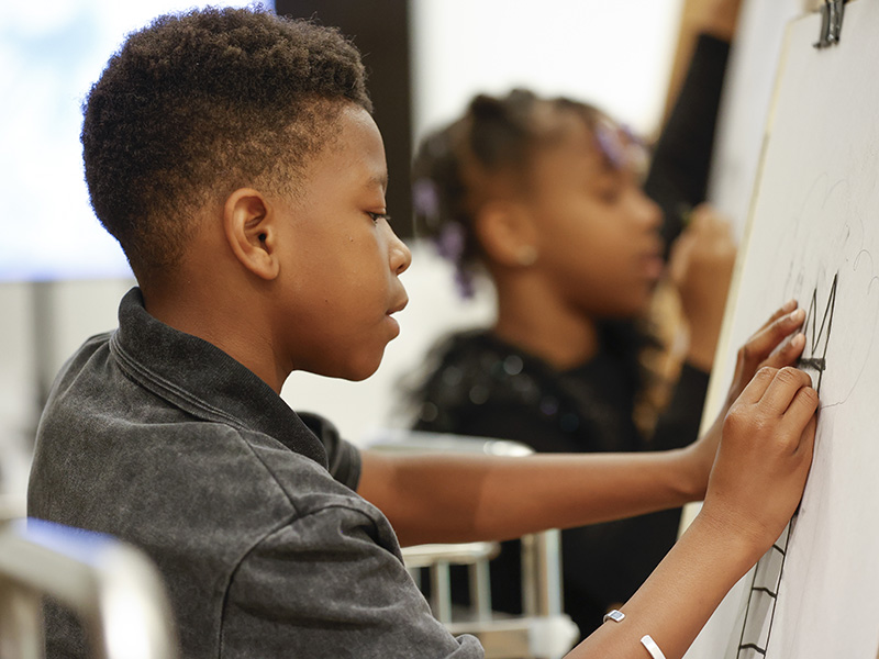 Photo of a young boy drawing on a sheet of paper set on an art easel.