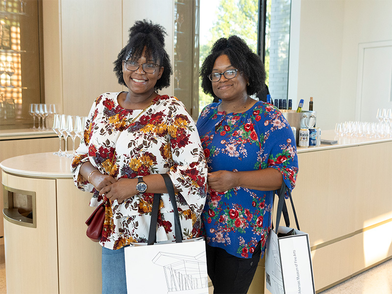 Photo of two women smiling and holding AMFA shopping bags in front of a bar.