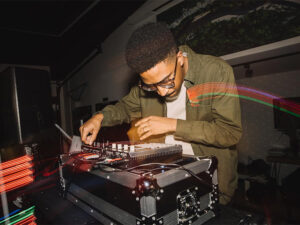Photo of a male DJ wearing a green jacket and glasses mixing music on a soundboard set on top of a music case.