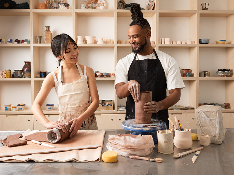 Photo of a woman and a man standing and hand rolling pieces of brown clay on a work surface in a ceramics studio.