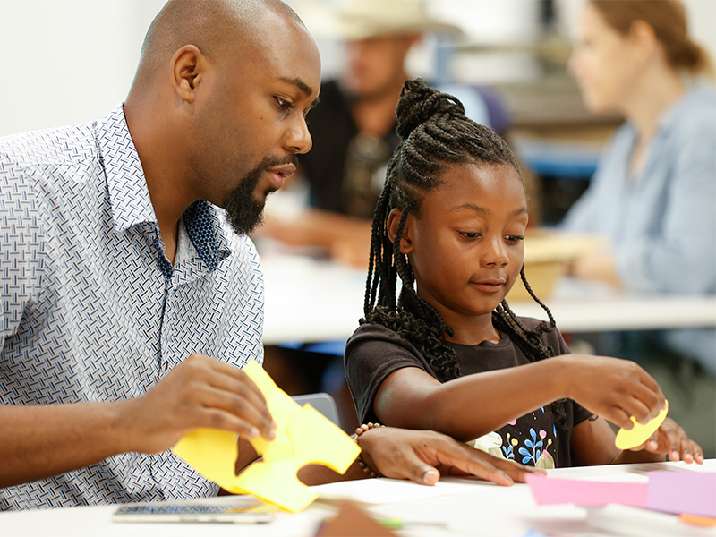 Photo of a father and daughter assembling cut paper shapes in an art studio.