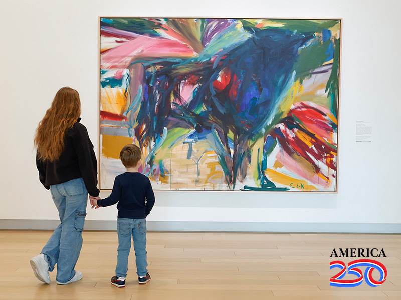 Photo of a girl and a young boy holding hands while standing in front of a large, colorful abstract painting of a bull.