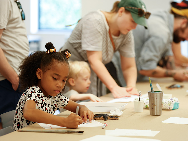 Photo of a young girl drawing on paper on a table in an art studio. Other children and adults are doing the same in the background.