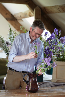 Photo of a man with short gray hair wearing glasses, a light blue button down shirt, and khakis arranging flowers in a vase on a wooden table.
