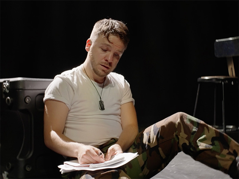 Photo of a male actor wearing camouflage pants and a white shirt sitting on a dark stage and writing in a notebook.