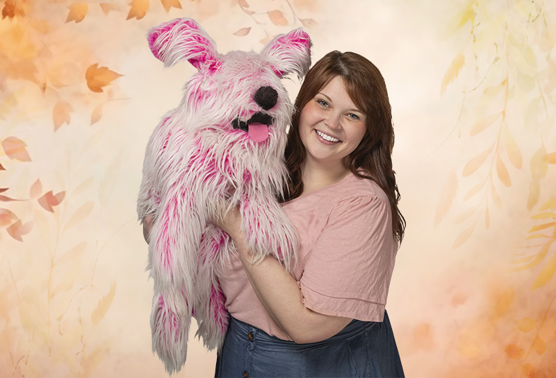 Photo of a woman wearing a pink shirt holding a pink dog puppet in front of a leafy background.