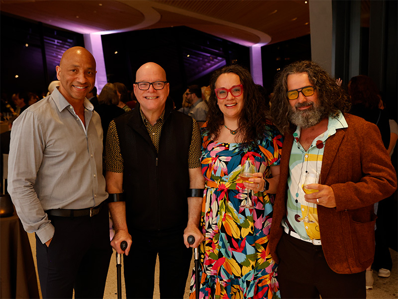 Photo of three men and one woman posed smiling while holding drinks at a party.