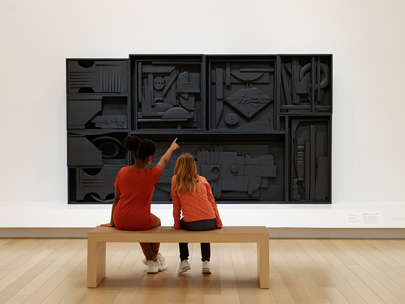 Photo of two young girls sitting on a bench in front of a sculpture by Louise Nevelson. The girl on the left is pointing to the sculpture.