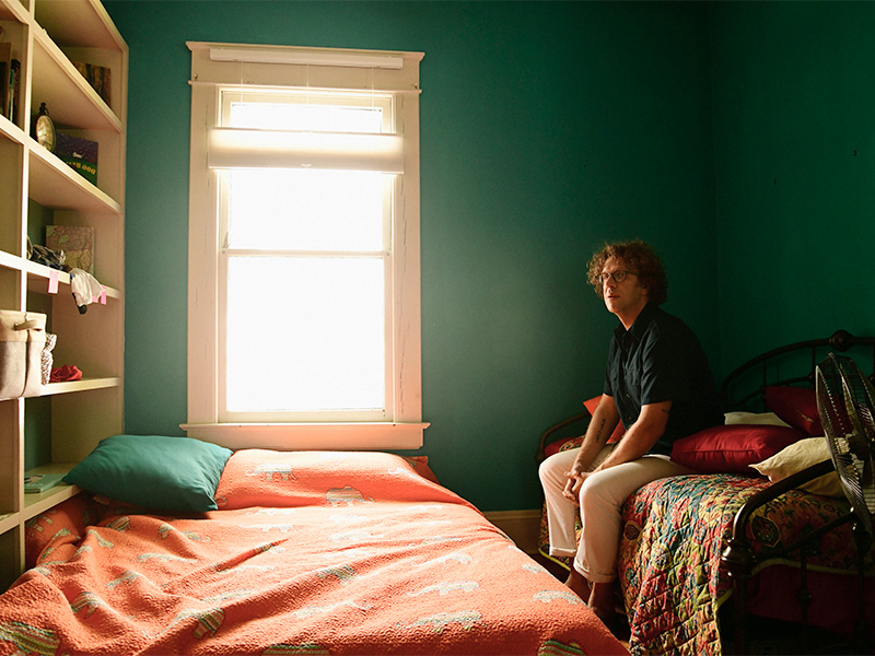 Photo of a man with short, curly brown hair and glasses sitting on a bed next to another bed in a room with book shelves and a window.