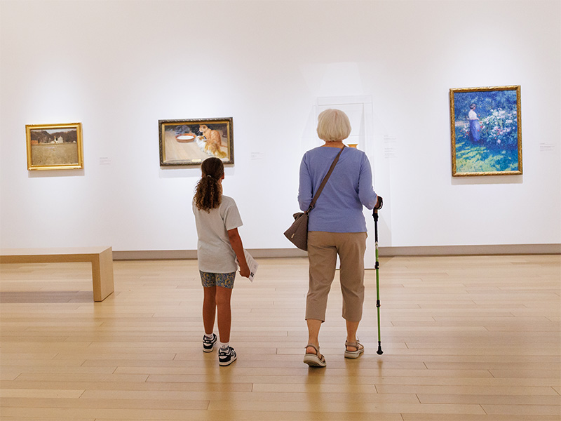 Photo of a young girl and elderly woman walking with a cane looking at art in an art gallery.