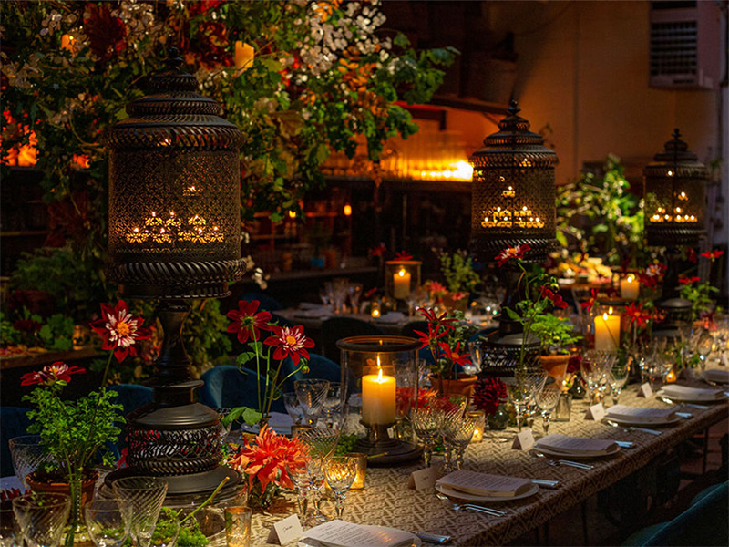 Photo of a long table set with plates, silverware, and glasses next to large floral arrangements and lanterns holding candles.