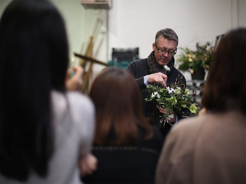 Photo of a man with short gray hair wearing glasses and a dark sweater and scarf arranging flowers in front of a group of people.