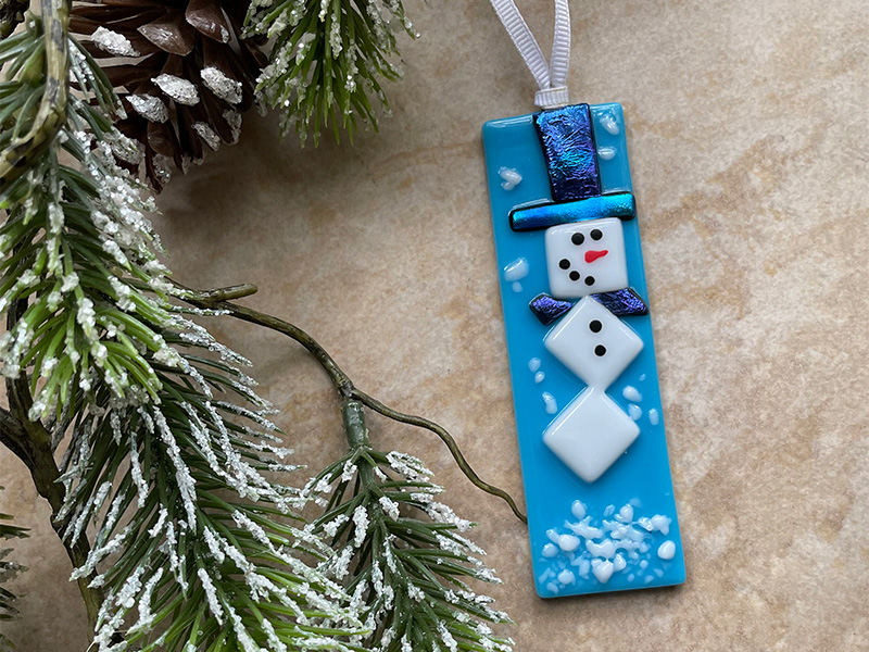 Photo of a flat fused glass snowman ornament laying on a tabletop next to a pine tree limb.