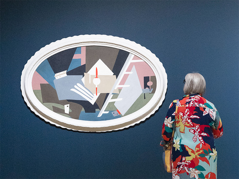 Photo of a woman wearing a floral dress standing in front of an abstract painting on a dark blue wall in an art gallery.