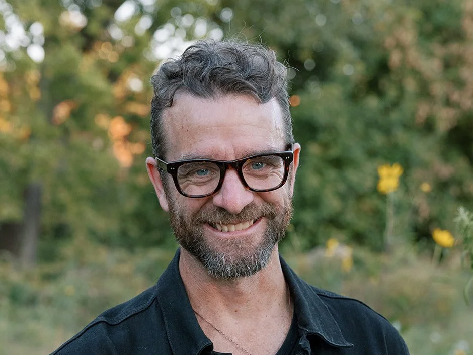 Headshot of a man wearing dark rimmed glasses and smiling while standing in front of a row of trees.